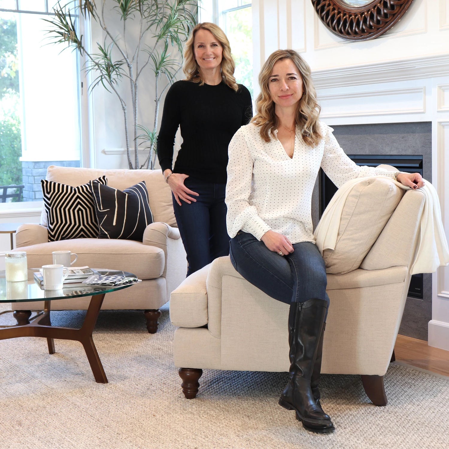 two blond women, one seated the other standing, both wearing jeans. Seated lady wearing white blouse with small black spots standing women wearing black long sleeve top.