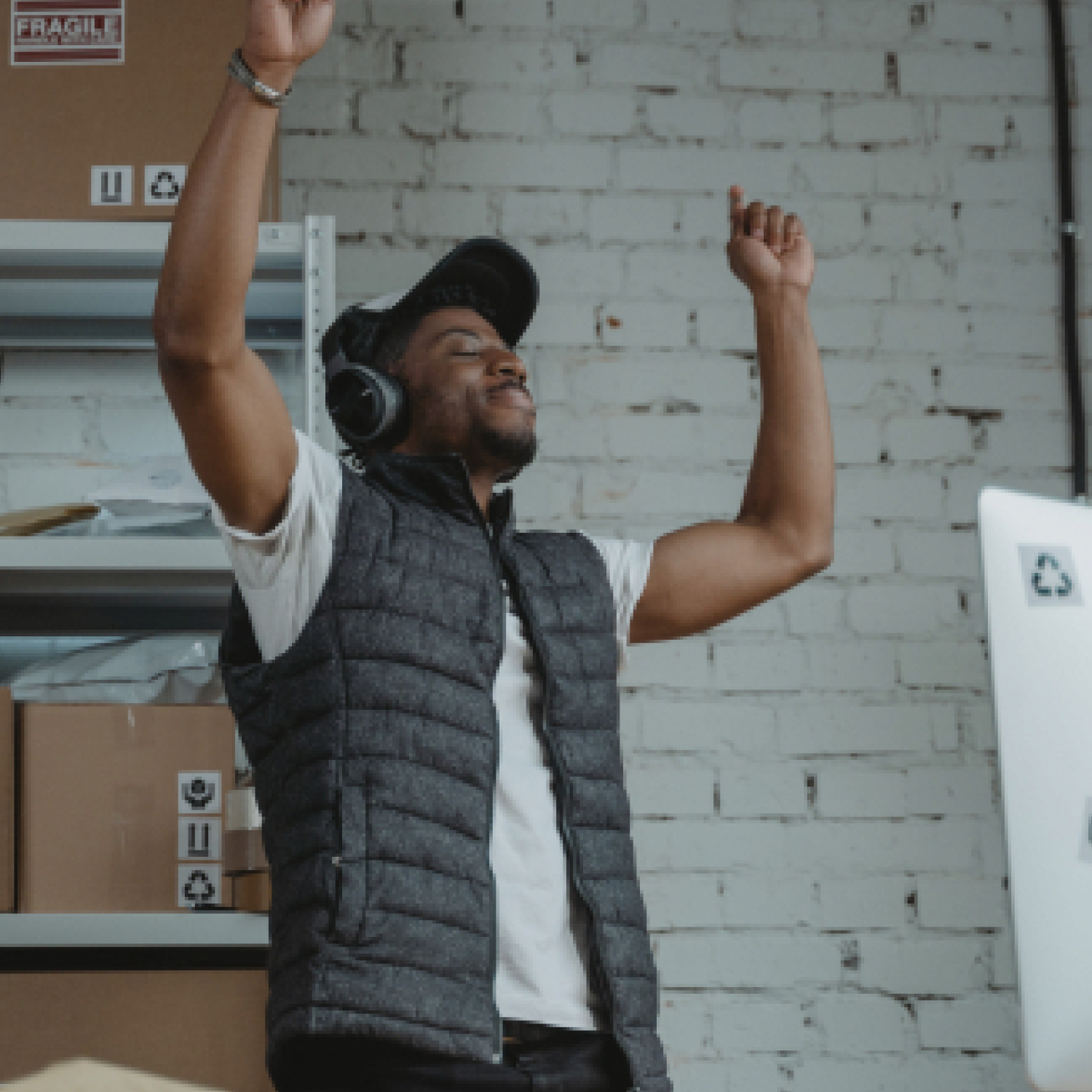 a man wearing headphones and an insulated vest in a stock room with his eyes closed and arms in the air in a dancing or celebrating gesture. 