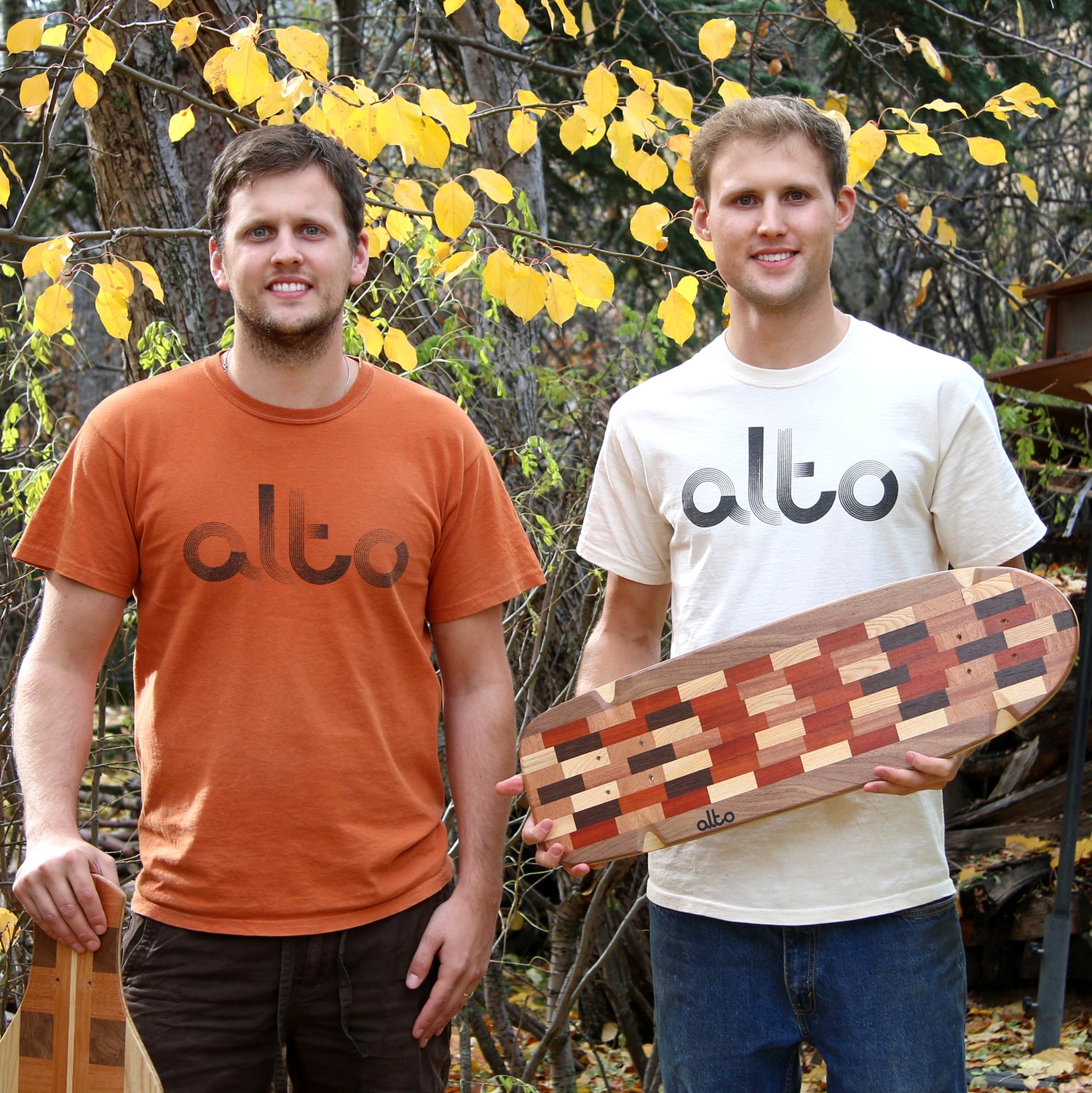 Two men standing with logo thirts. One in orange and one in white. The man on the right is holding a wooden skateboard
